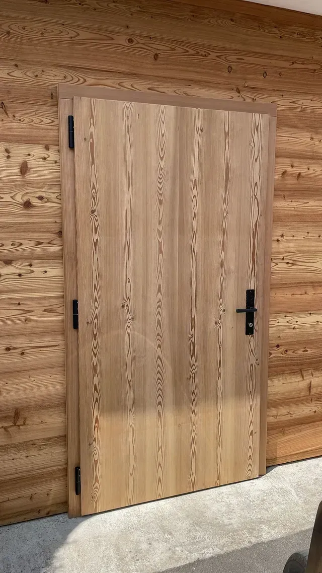 Porte en bois clair fermée dans un mur de cabane en rondins, avec poignée et charnières noires.