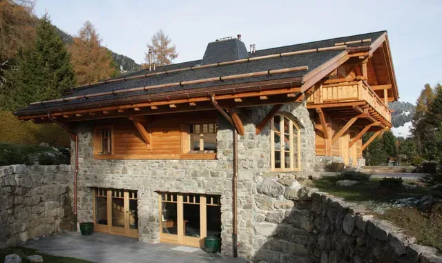 Chalet en pierre et en bois avec de grandes fenêtres et un toit en pente dans un cadre montagneux boisé