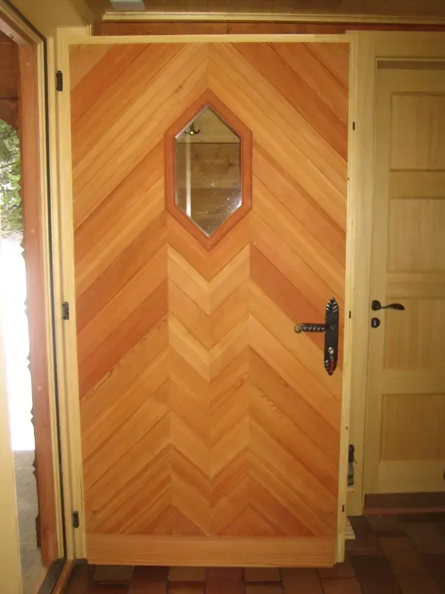 Porte d'entrée en bois à panneaux en chevrons et petite fenêtre en losange, légèrement ouverte vers l'intérieur.