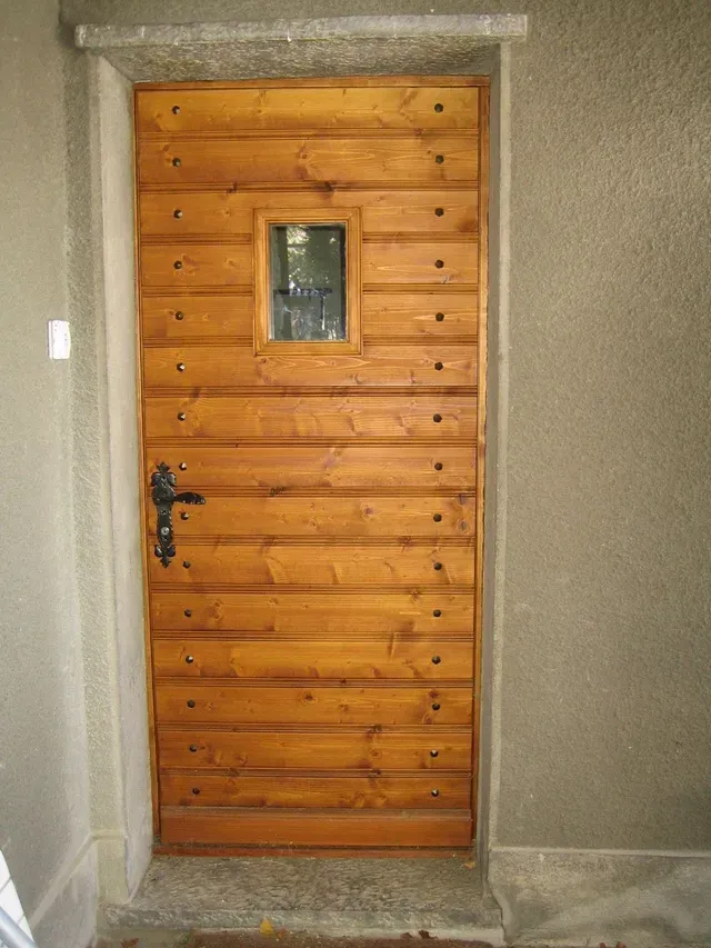 Porte d'entrée en bois avec une petite fenêtre, encastrée dans un mur en stuc gris.
