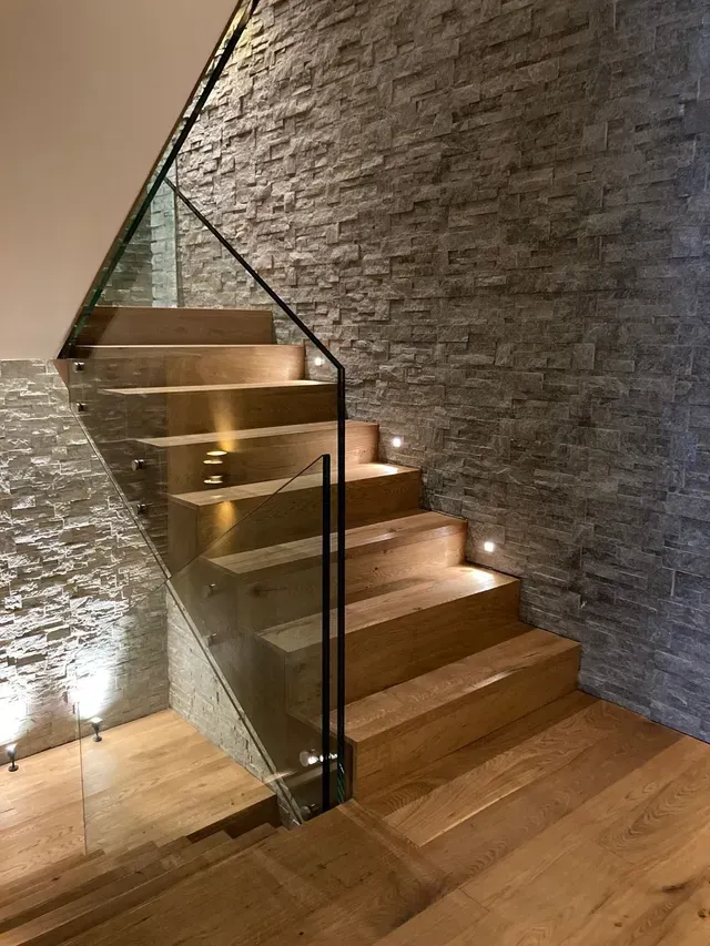 Escalier moderne en bois avec rampe en verre et mur en pierre, éclairé par de petits spots encastrés dans les marches.