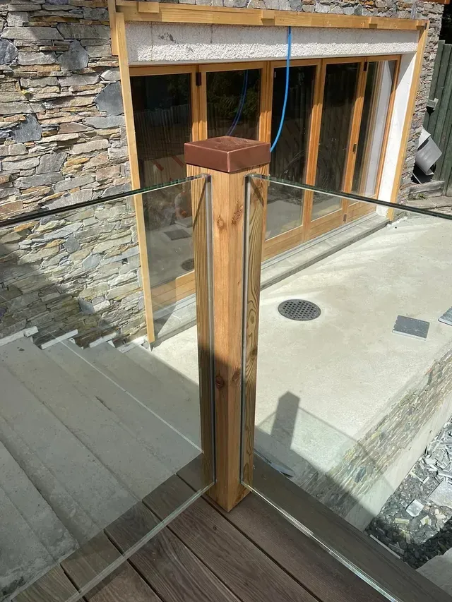 Poteau de garde-corps en bois et verre sur une terrasse jouxtant une maison en pierre.