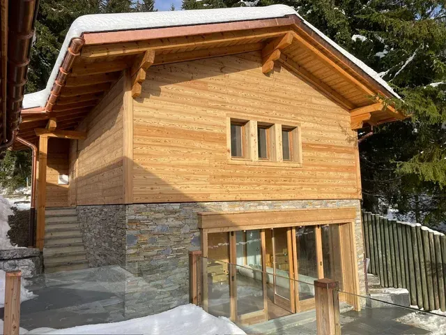 Maison de style chalet en bois avec base en pierre et grandes portes vitrées dans un cadre forestier enneigé