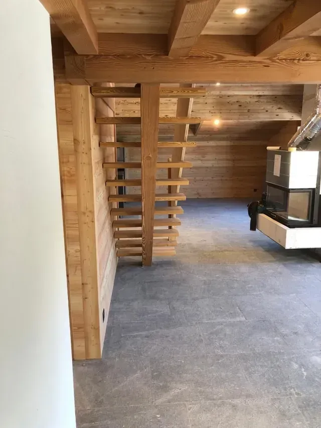 Intérieur de loft en bois avec escalier escamotable, poutres apparentes, moquette grise et banquette près de la fenêtre.