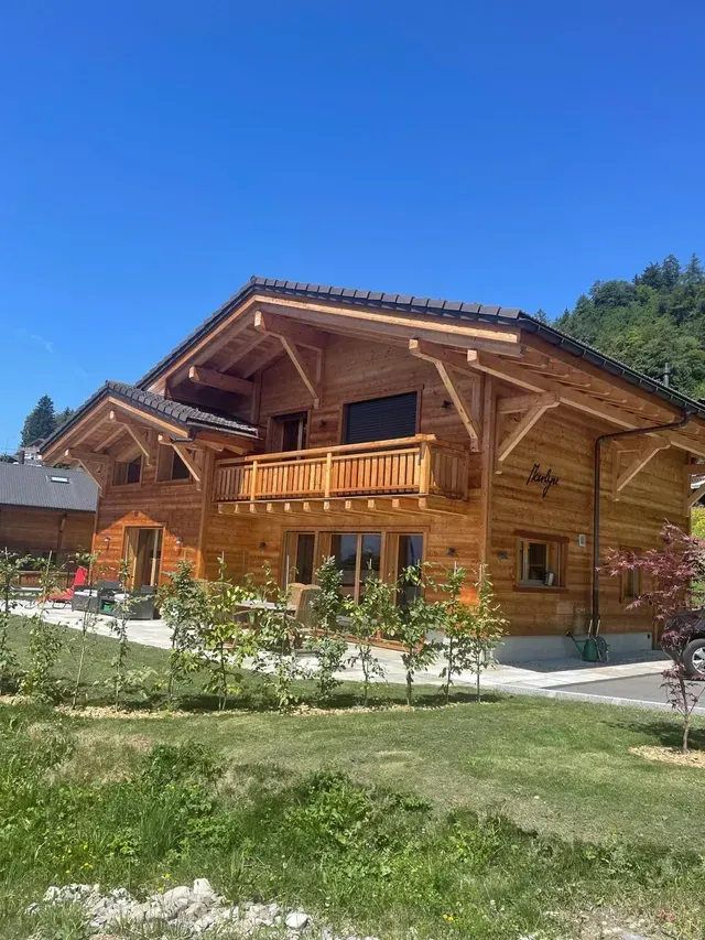 Maison de style chalet en bois avec balcon et jardin sous un ciel bleu clair