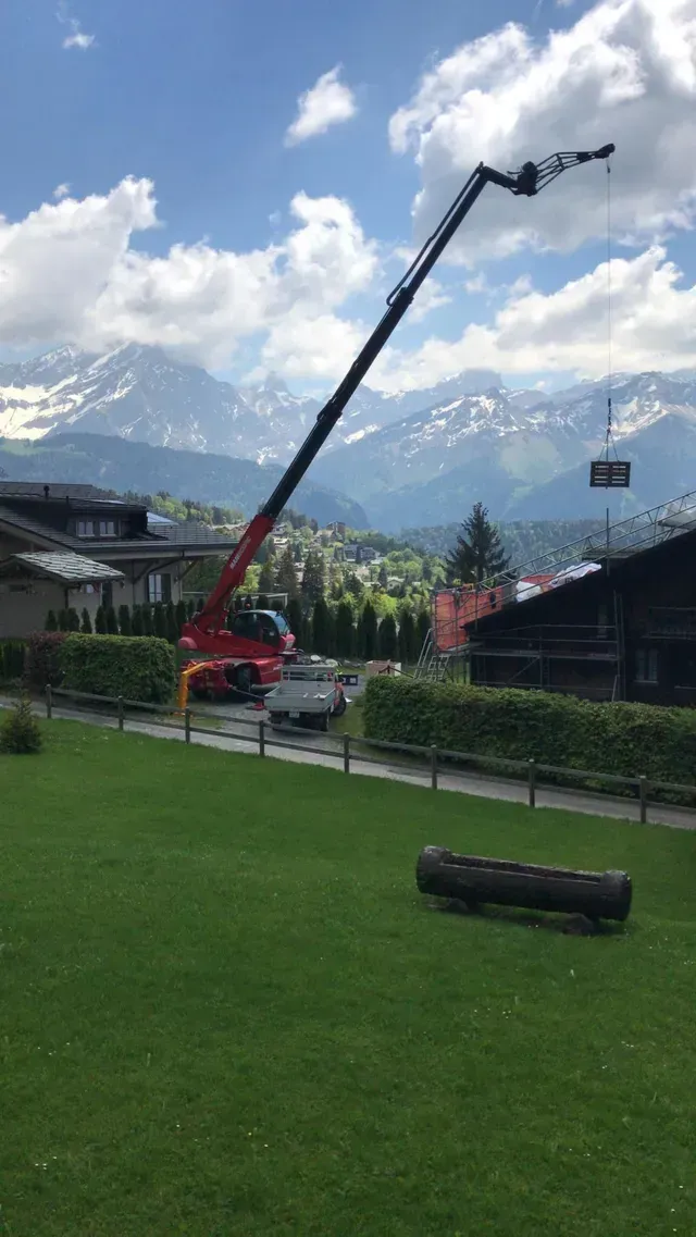 Une grue rouge soulève une télécabine près de bâtiments alpins, avec des montagnes et des nuages ​​en arrière-plan.