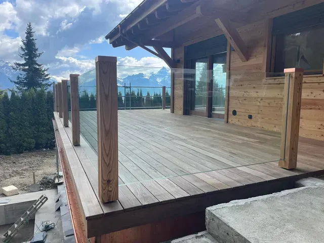Terrasse en bois jouxtant un chalet avec vue sur la montagne et balustrade en verre au coucher du soleil.
