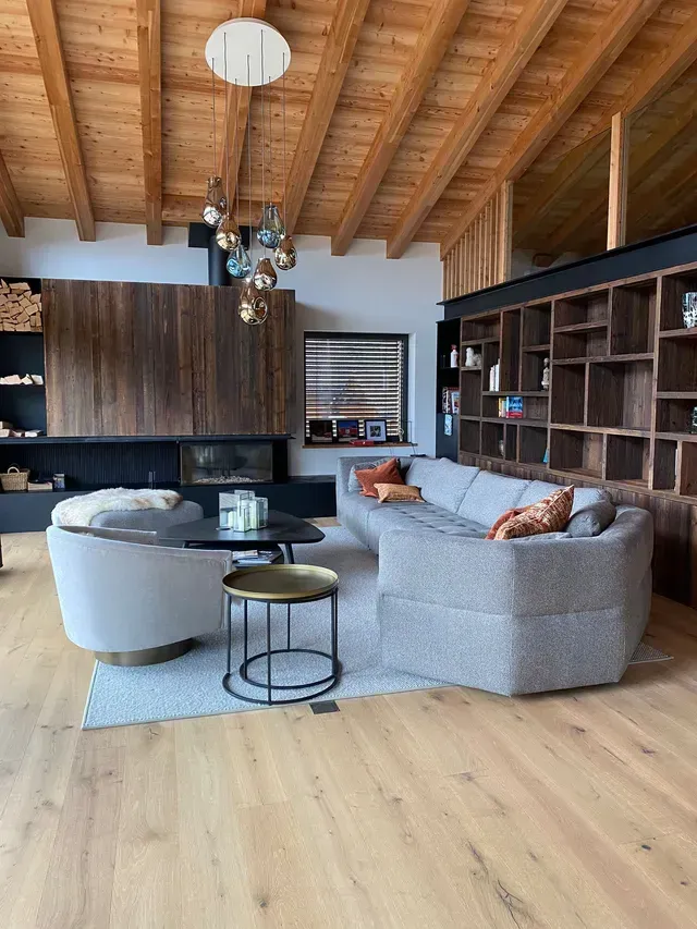 Salon moderne avec plafond en bois, canapé d'angle gris, fauteuils ronds et étagères intégrées.