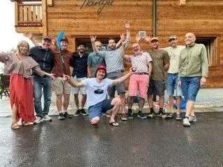 Un groupe de personnes pose en extérieur devant un bâtiment en bois, souriant et levant les bras.