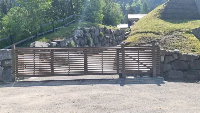 Portail en bois à claire-voie barrant une route pavée, longeant un canal bordé de pierres et un talus herbeux.
