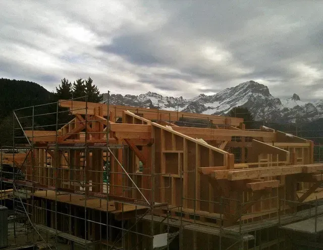 Maison à ossature bois en construction, avec des échafaudages, se détachant sur un fond de montagnes nuageuses.
