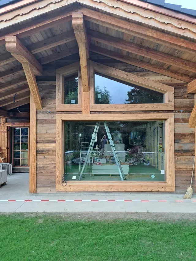 Façade de maison en bois avec une grande fenêtre en façade, des poutres apparentes et une échelle à l'intérieur
