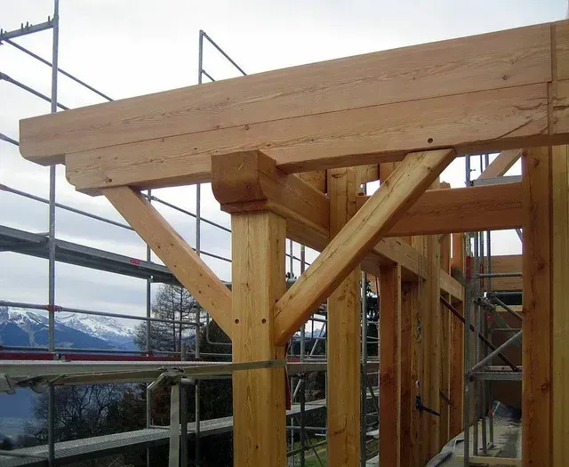 Charpente de toit en bois en construction sur un échafaudage, avec des montagnes visibles en arrière-plan.