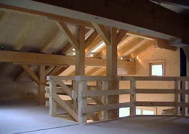 Grenier aménagé en bois avec poutres apparentes, balustrade et une petite fenêtre éclairée d'une lumière chaude.