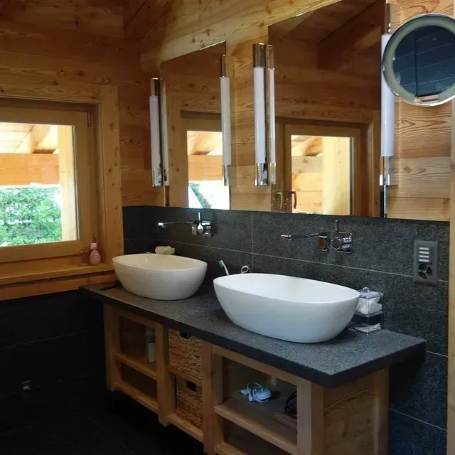 Salle de bains lambrissée avec deux vasques, comptoir en pierre foncée et miroir rond près des fenêtres.