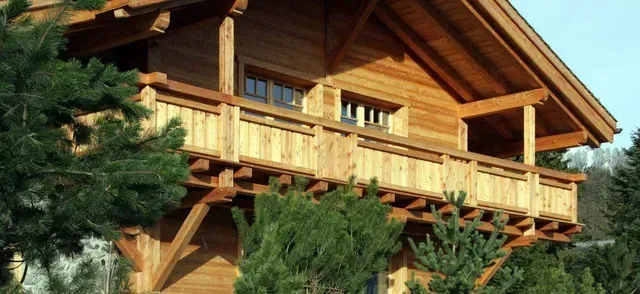 Chalet en bois avec balcon, partiellement caché par des arbres verts, baigné d'un chaud soleil