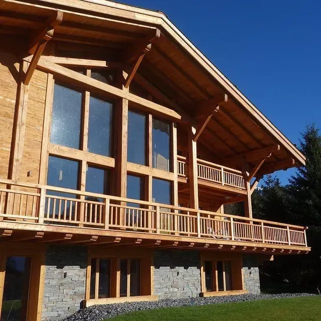 Cabane en bois avec de grandes baies vitrées, un socle en pierre et un balcon panoramique sous un ciel bleu azur.