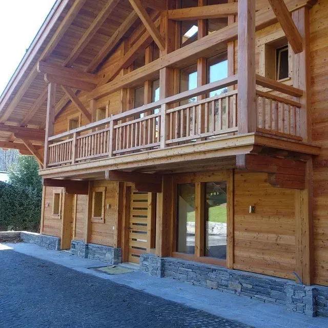 Chalet en bois avec balcon couvert et soubassement en pierre, à côté d'une allée de gravier.