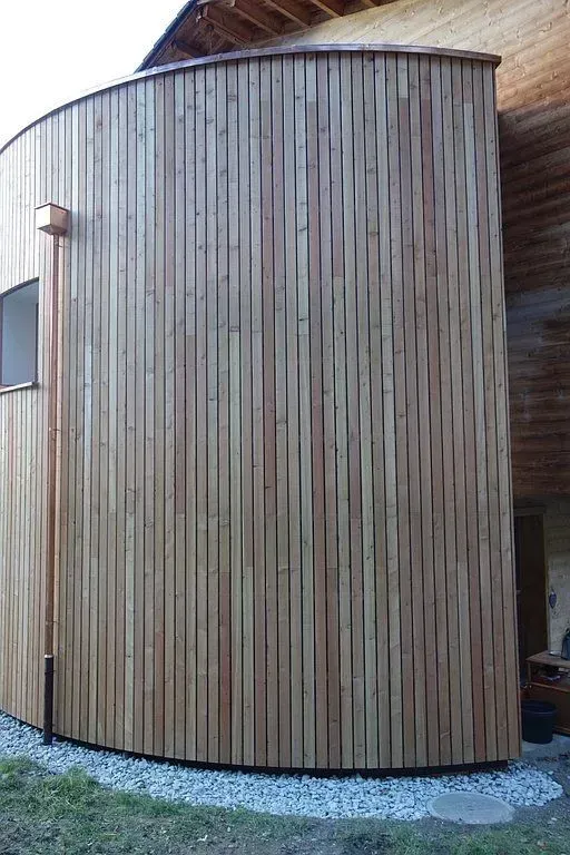 Façade en bois courbée avec des lattes verticales et un chemin de gravier à la base