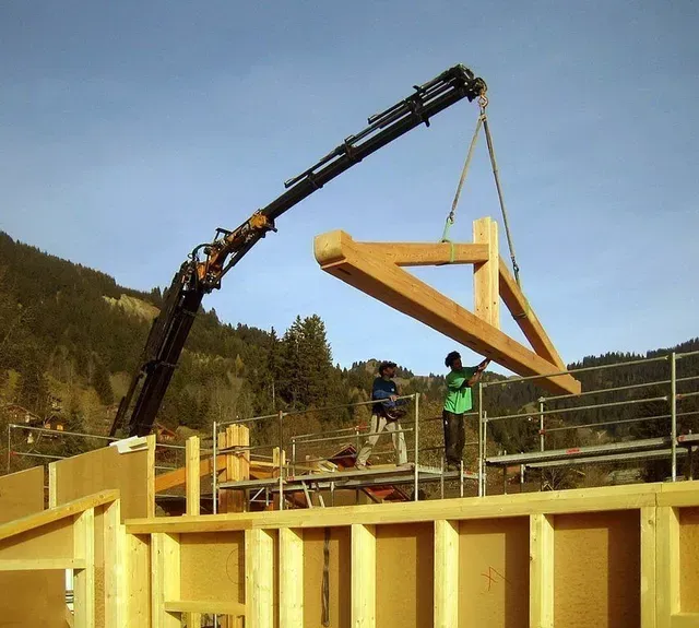 Une grue soulève une ferme de toit en bois sur un chantier de construction de maison, des ouvriers se trouvant en dessous.