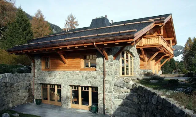Chalet en pierre et en bois, avec un toit en pente et de grandes fenêtres, niché au milieu des rochers et des arbres.