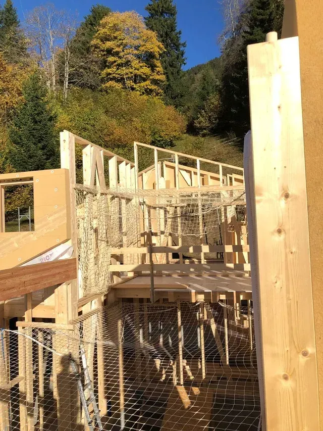 La structure en bois d'un bâtiment est en construction sur un versant ensoleillé, avec des arbres en arrière-plan.