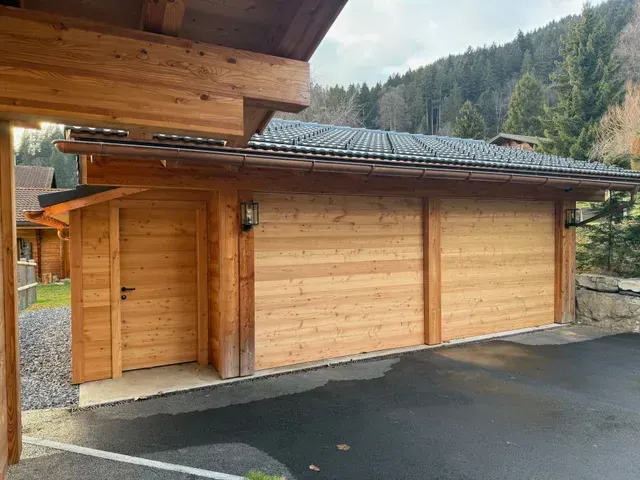 Cabane en bois de style chalet avec un toit sombre, située à côté d'une allée humide et d'une colline boisée.