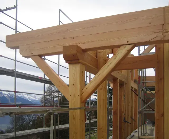 Charpente de toit en bois en construction, avec échafaudages et montagnes enneigées en arrière-plan.