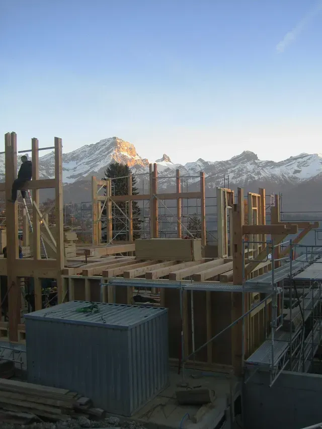 Chantier de construction avec charpente en bois et échafaudages se détachant sur des montagnes enneigées au lever du soleil