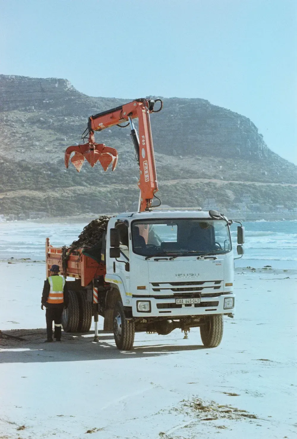 Camión con grúa recogiendo escombros en una playa; trabajador con chaleco. Montañas al fondo.