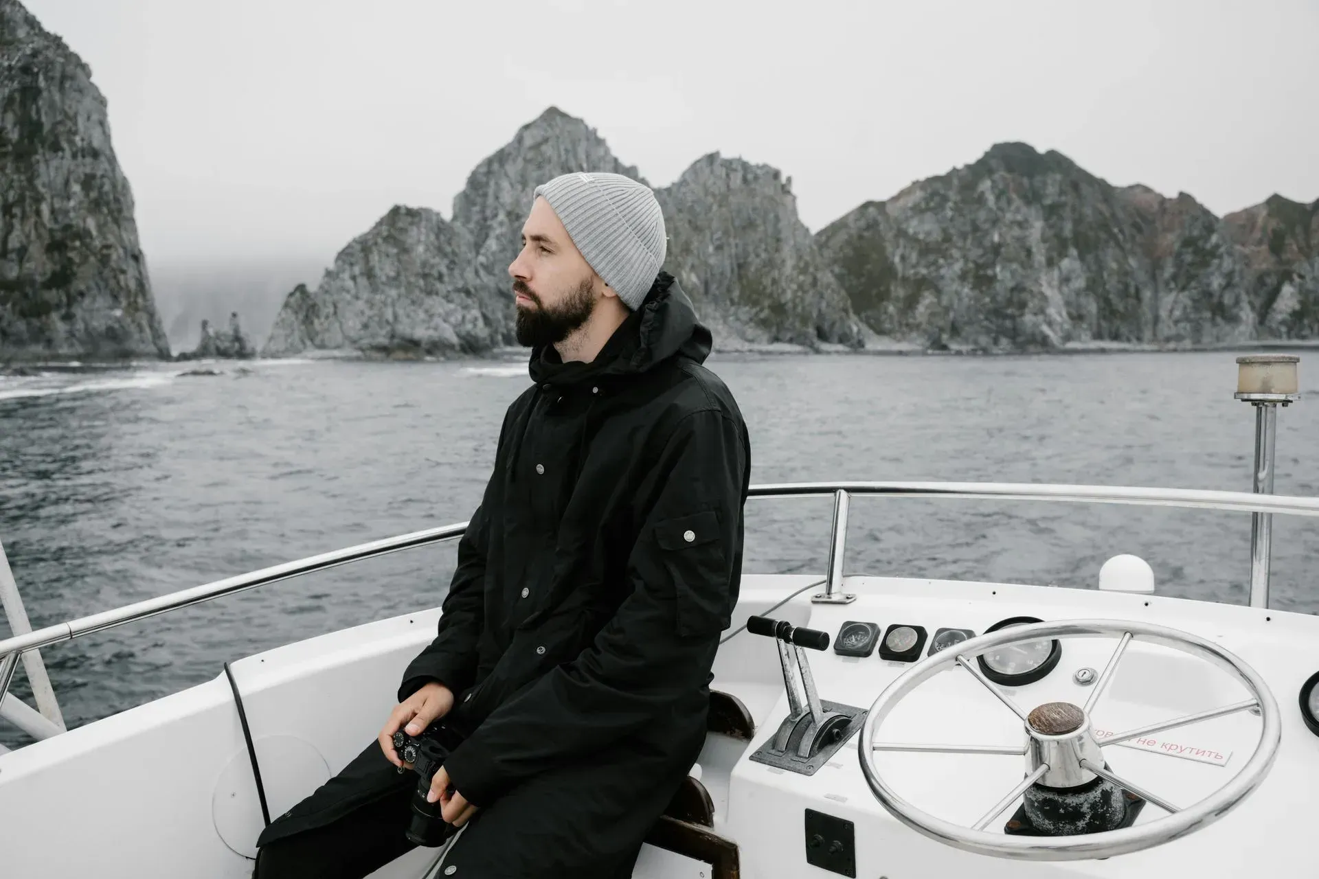 Hombre con gorro de lana y chaqueta negra en un barco, contemplando los acantilados rocosos y el mar.