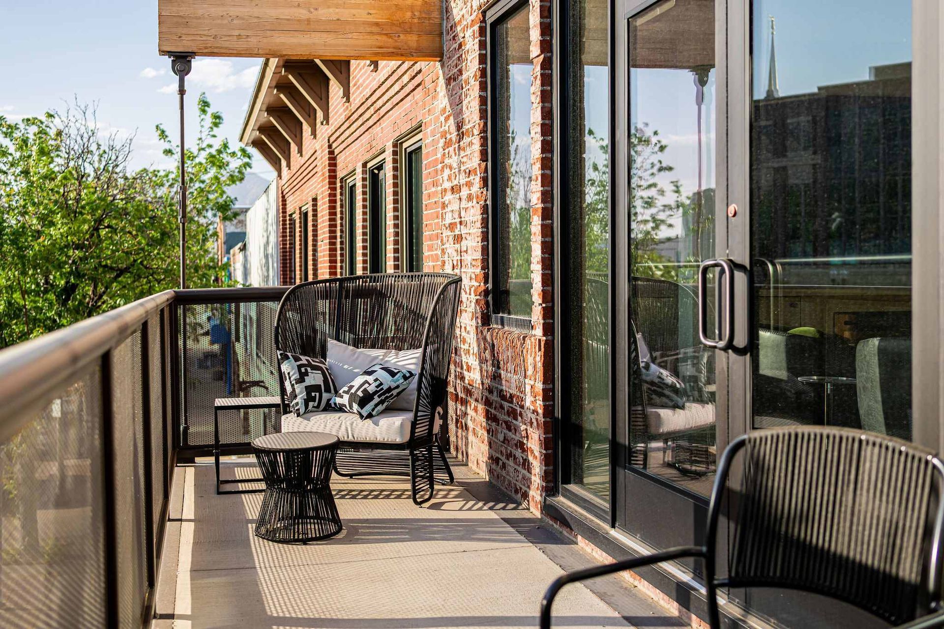 Balcony with wicker furniture, brick wall, glass doors, city view, sunny day.