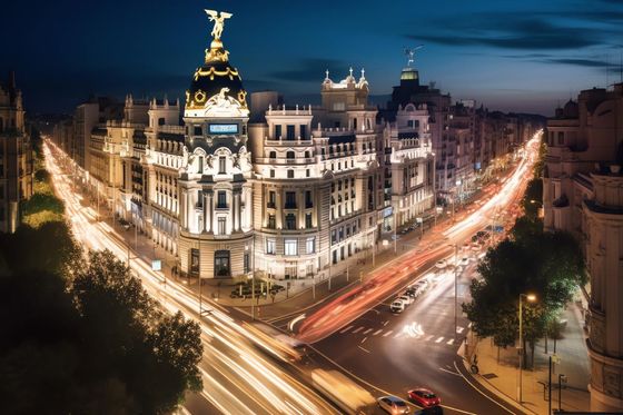 Vista nocturna de la Gran Vía de Madrid, larga exposición con luces brillantes de vehículos en la carretera que convergen hacia un gran edificio ornamentado.