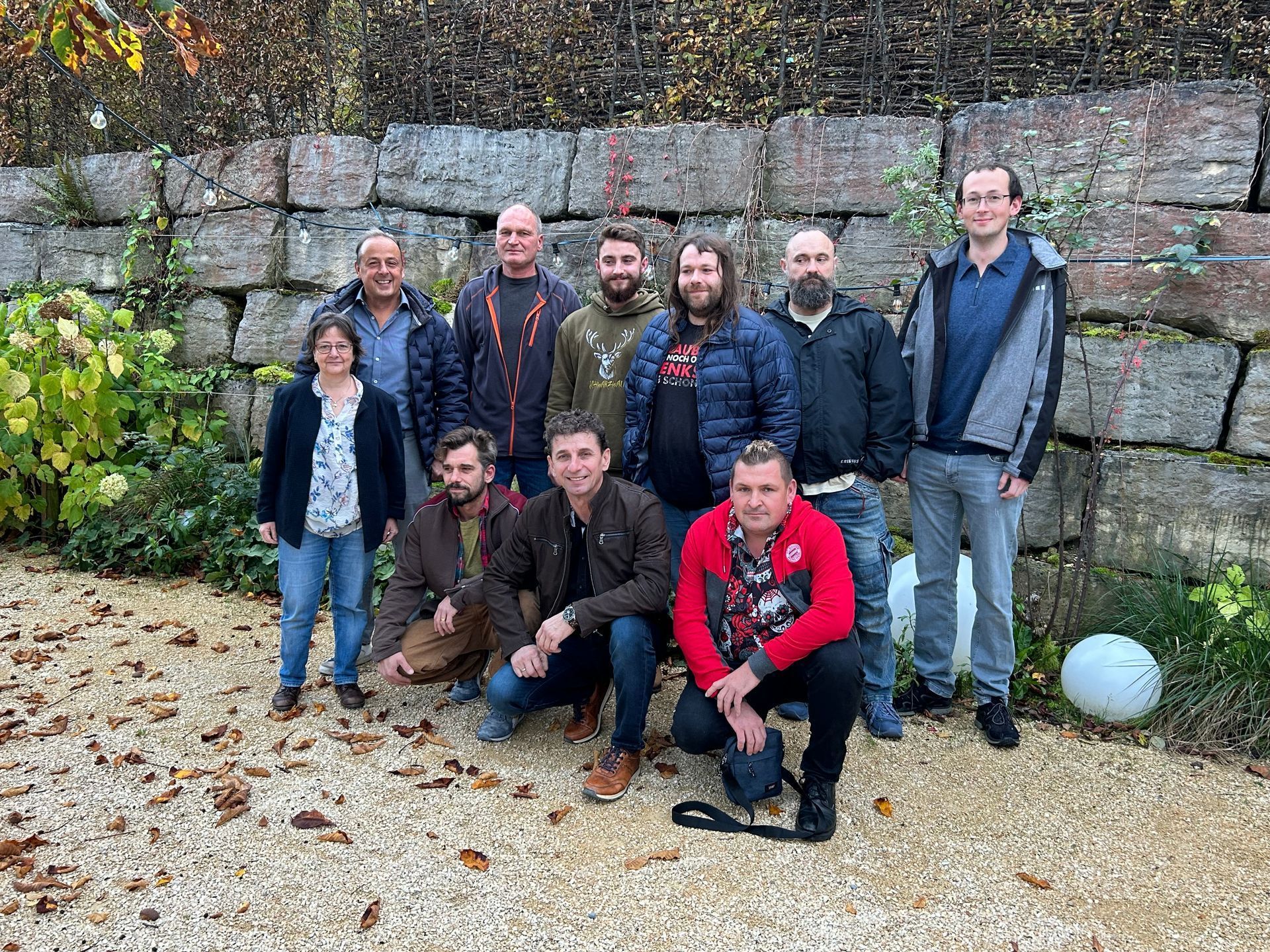 Eine Gruppe von Menschen posiert im Freien vor einer Steinmauer, eine Person kniet davor. Eine Gruppe von Menschen posiert im Freien vor einer Steinmauer, eine Person kniet davor.
