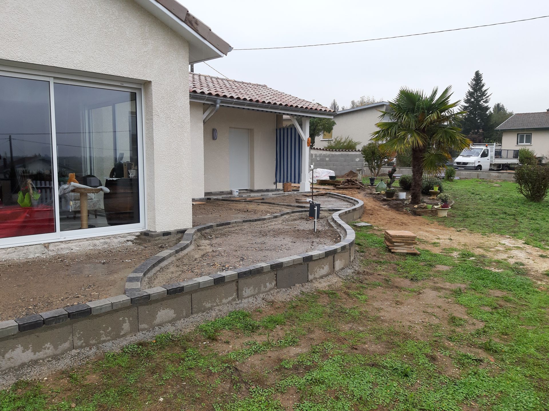 Terrasse de maison avec muret de soutènement incurvé, porte-fenêtre coulissante et jardin gazonné sous un ciel couvert.