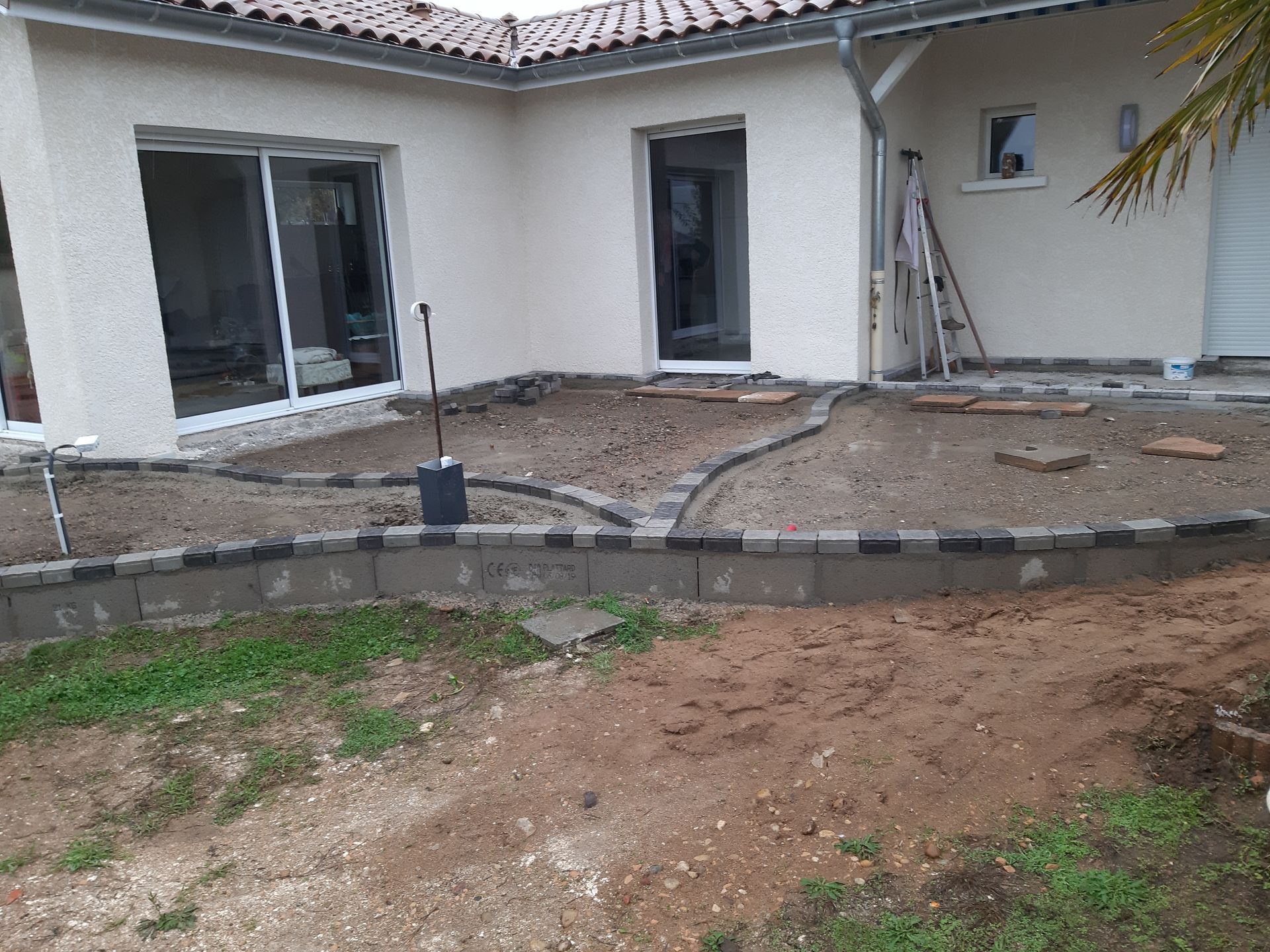 Terrasse en construction dans le jardin d'une maison blanche, avec des pavés, de la terre et des outils visibles.