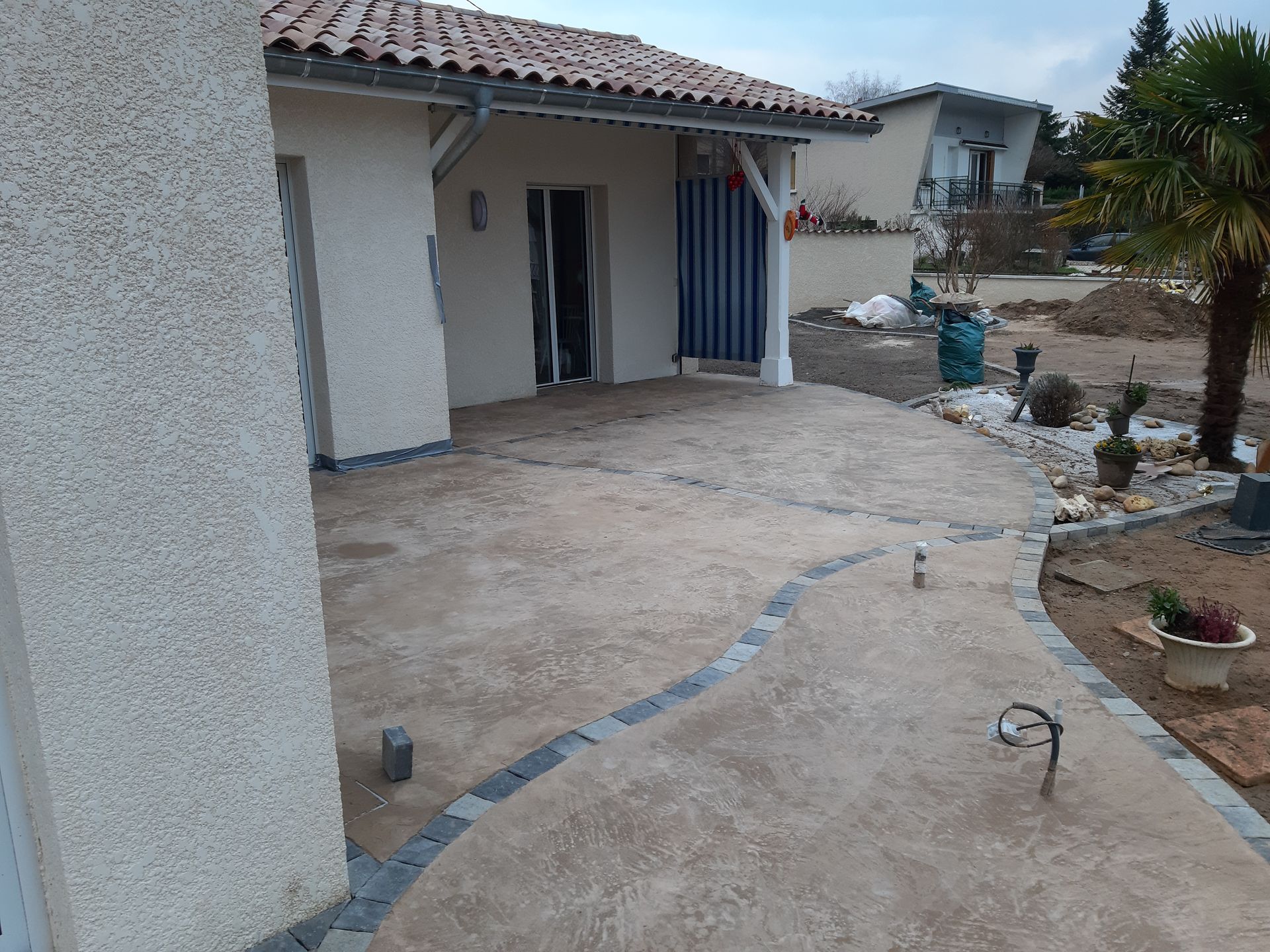 Terrasse en béton jouxtant une maison blanche au toit de tuiles et au portail bleu, bordant une cour en terre battue.