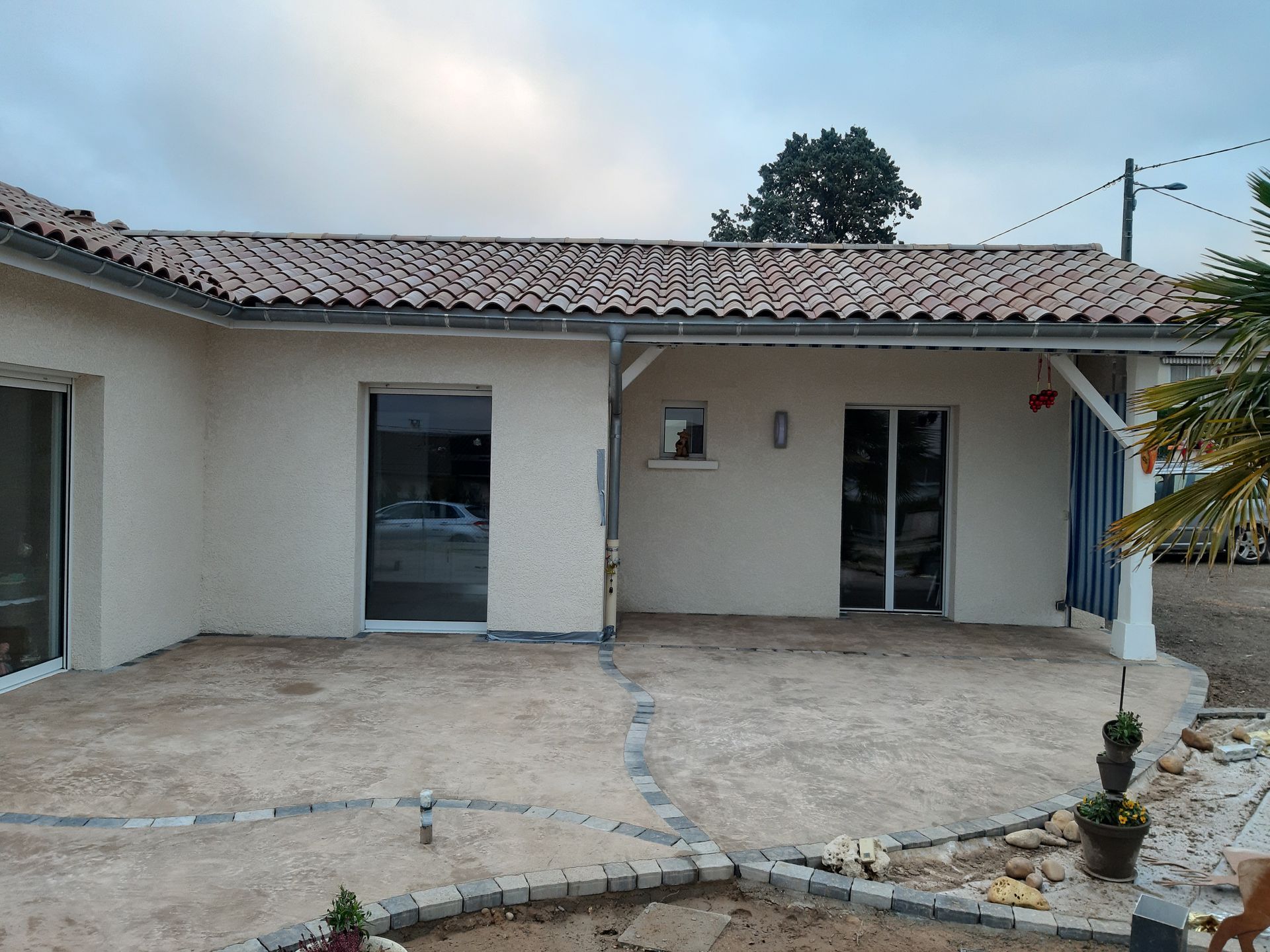 Patio d'une maison de plain-pied avec murs blancs, toit de tuiles et portes coulissantes en verre sous un ciel nuageux