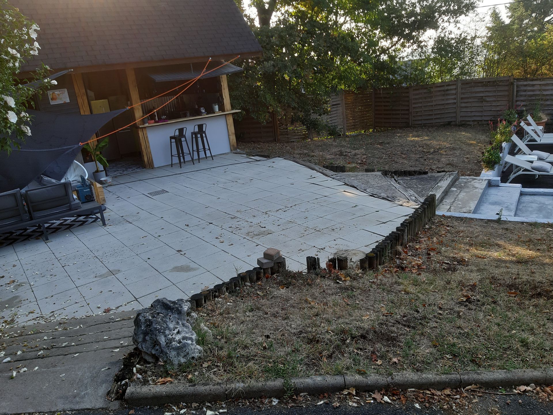 Terrasse arrière avec pavés gris, abri de jardin en bois à côté d'une zone de pelouse en pente