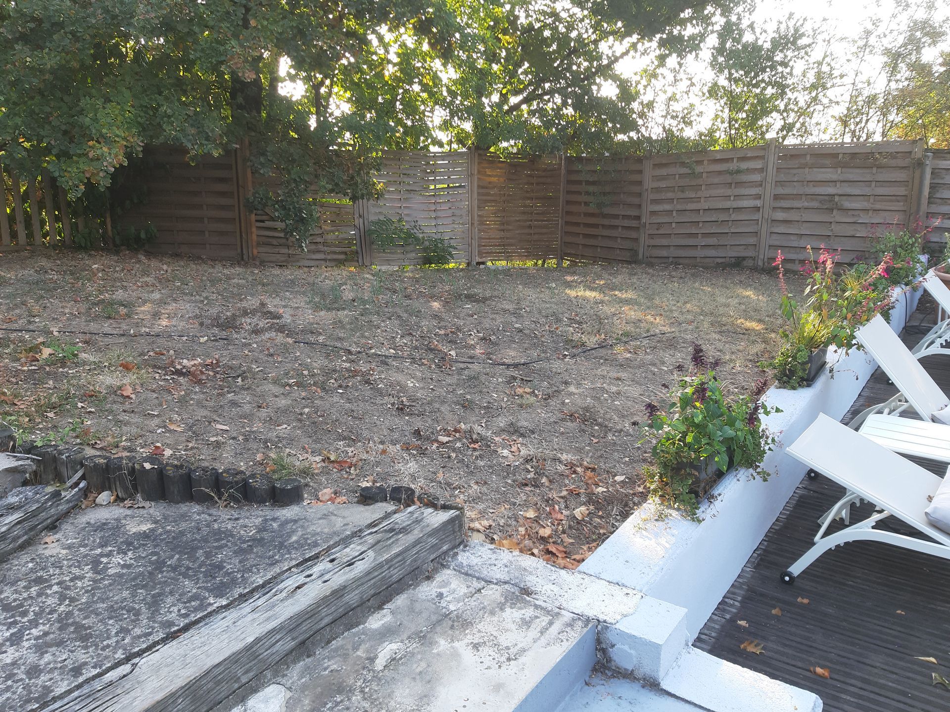 Terrasse arrière avec clôture en bois, arbres et chaises blanches à côté d'un jardin en terre battue.