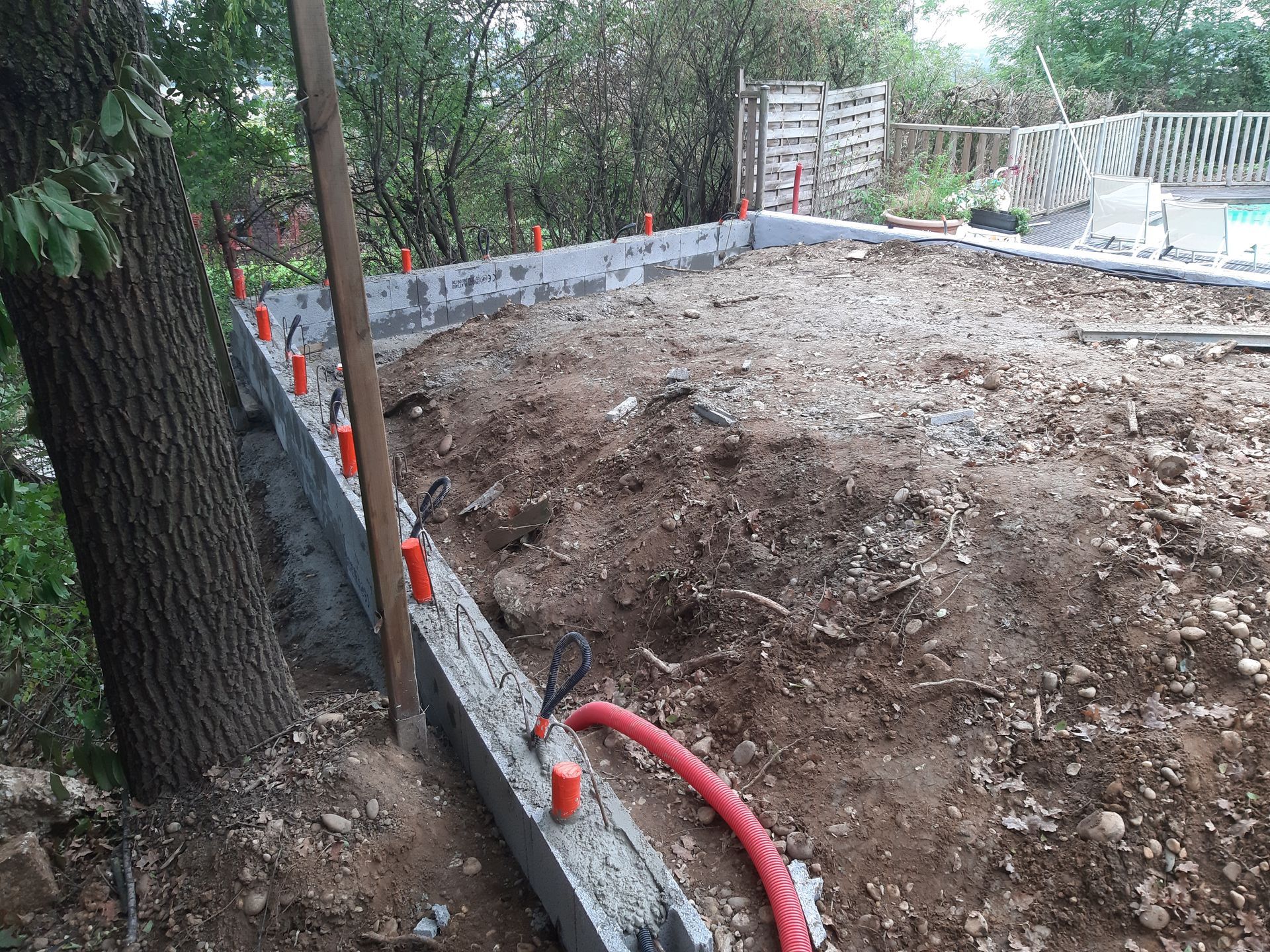 Chantier de construction avec terrain vague défriché, tuyau d'arrosage rouge et bordure en béton bas le long des arbres