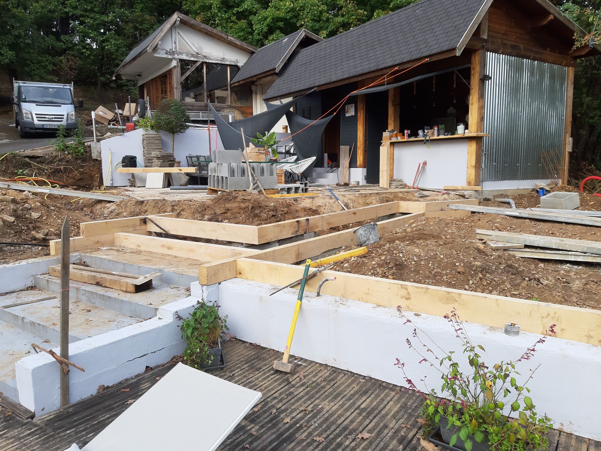 Chantier de construction avec les fondations d'une maison, une charpente en bois et un petit hangar en arrière-plan