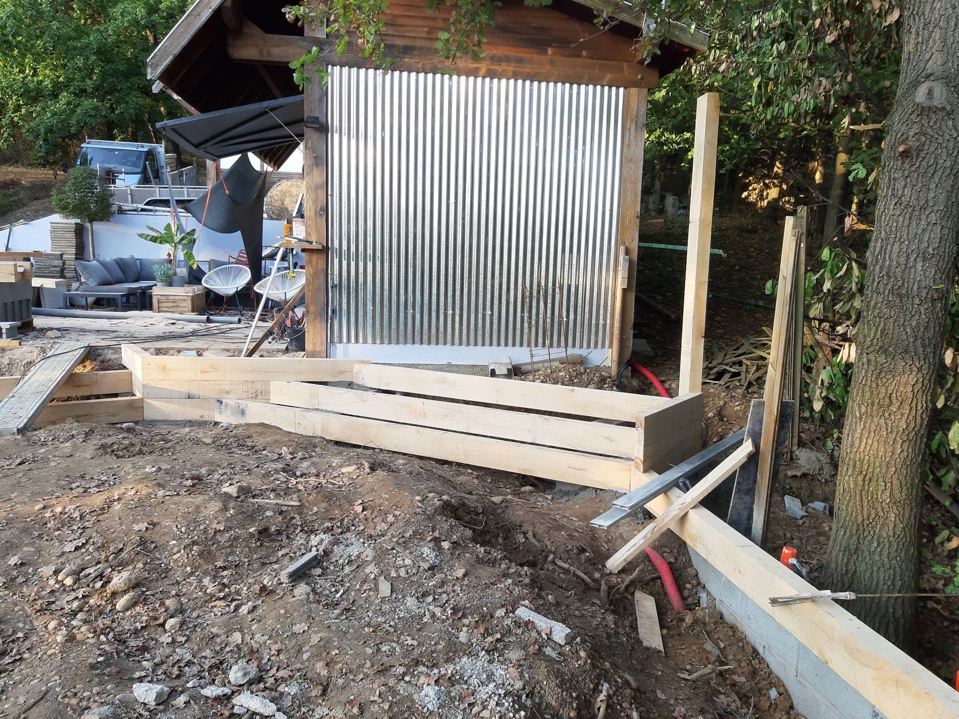 Zone de construction extérieure avec un abri en tôle ondulée, des marches en béton et des tuyaux apparents près d'un arbre