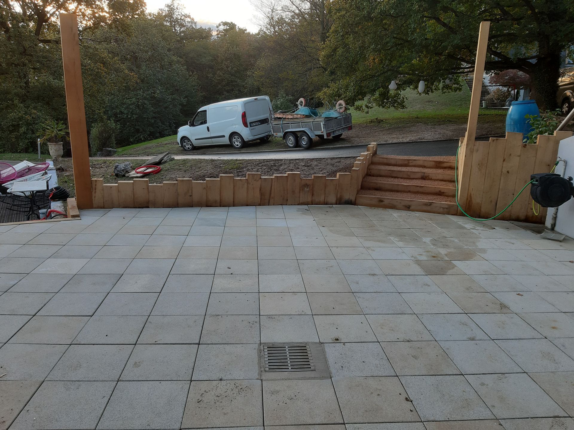 Terrasse en pierre avec un caniveau central, mur de soutènement en bois, et une camionnette et une remorque en arrière-plan.