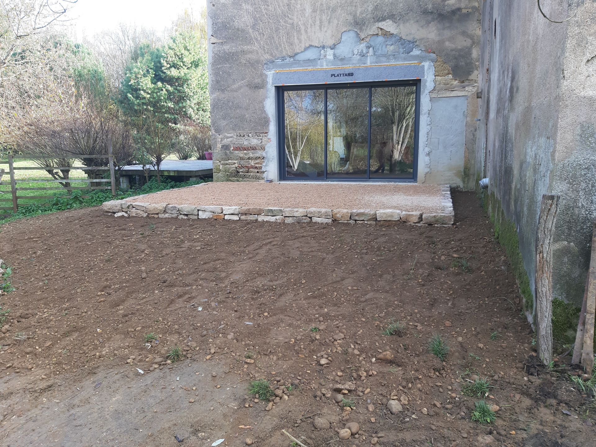 Terre nue jouxtant un bâtiment en pierre avec une baie vitrée et verdure en arrière-plan.