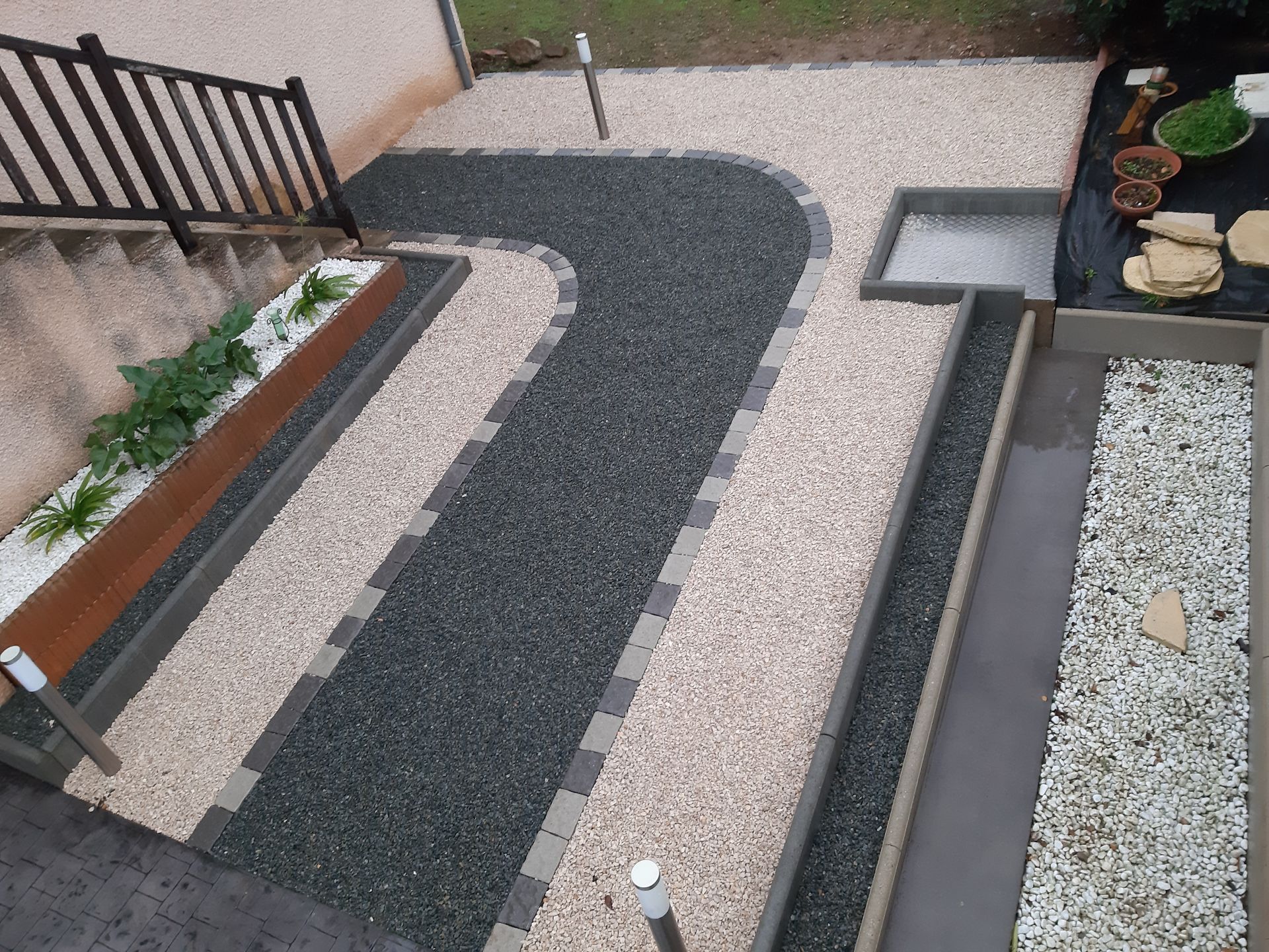 Allée sinueuse en gravier foncé bordée de pierres claires à côté d'une maison avec escalier.