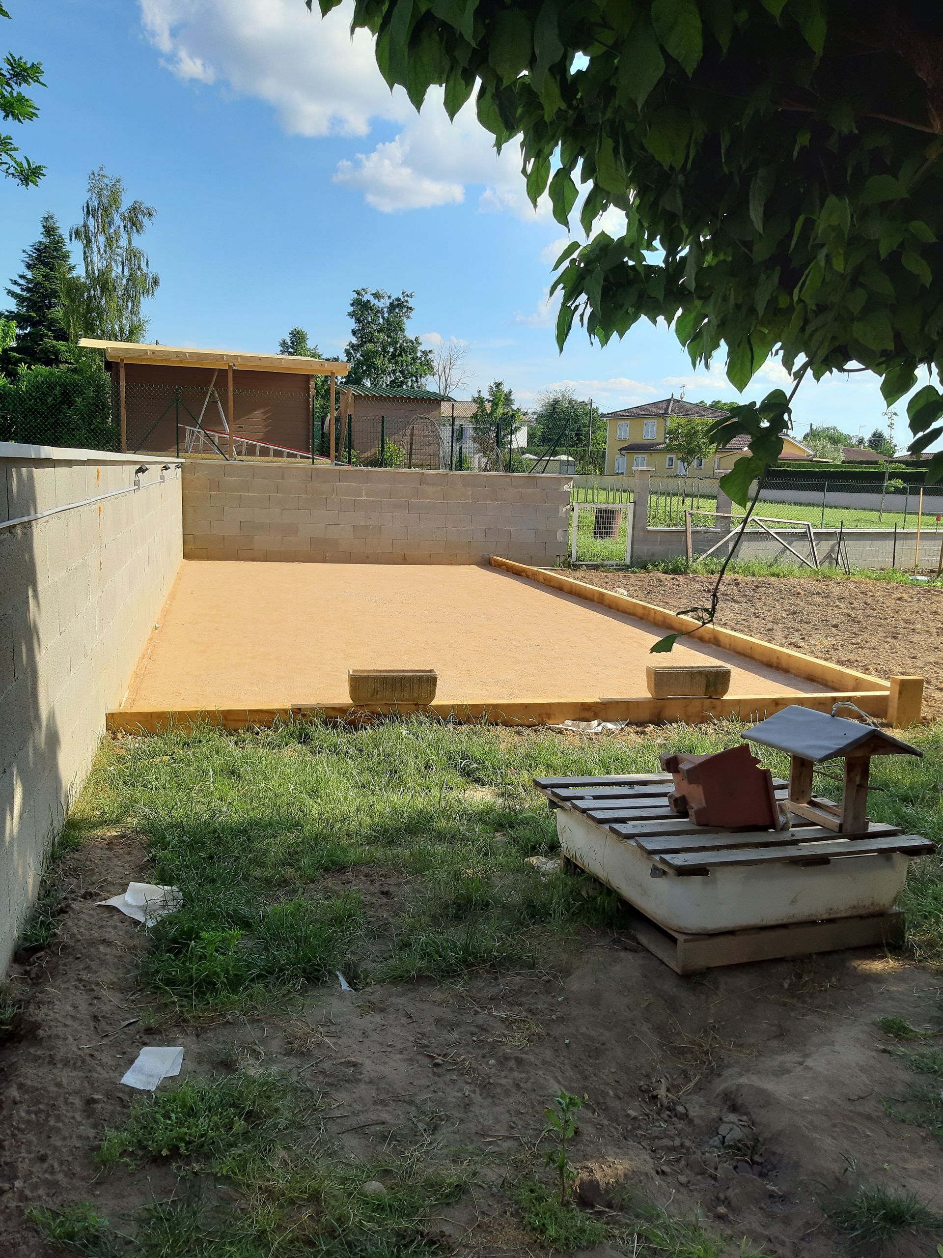 Jardin avec un mur beige, une pelouse et une petite table en bois sous les arbres et le ciel bleu.