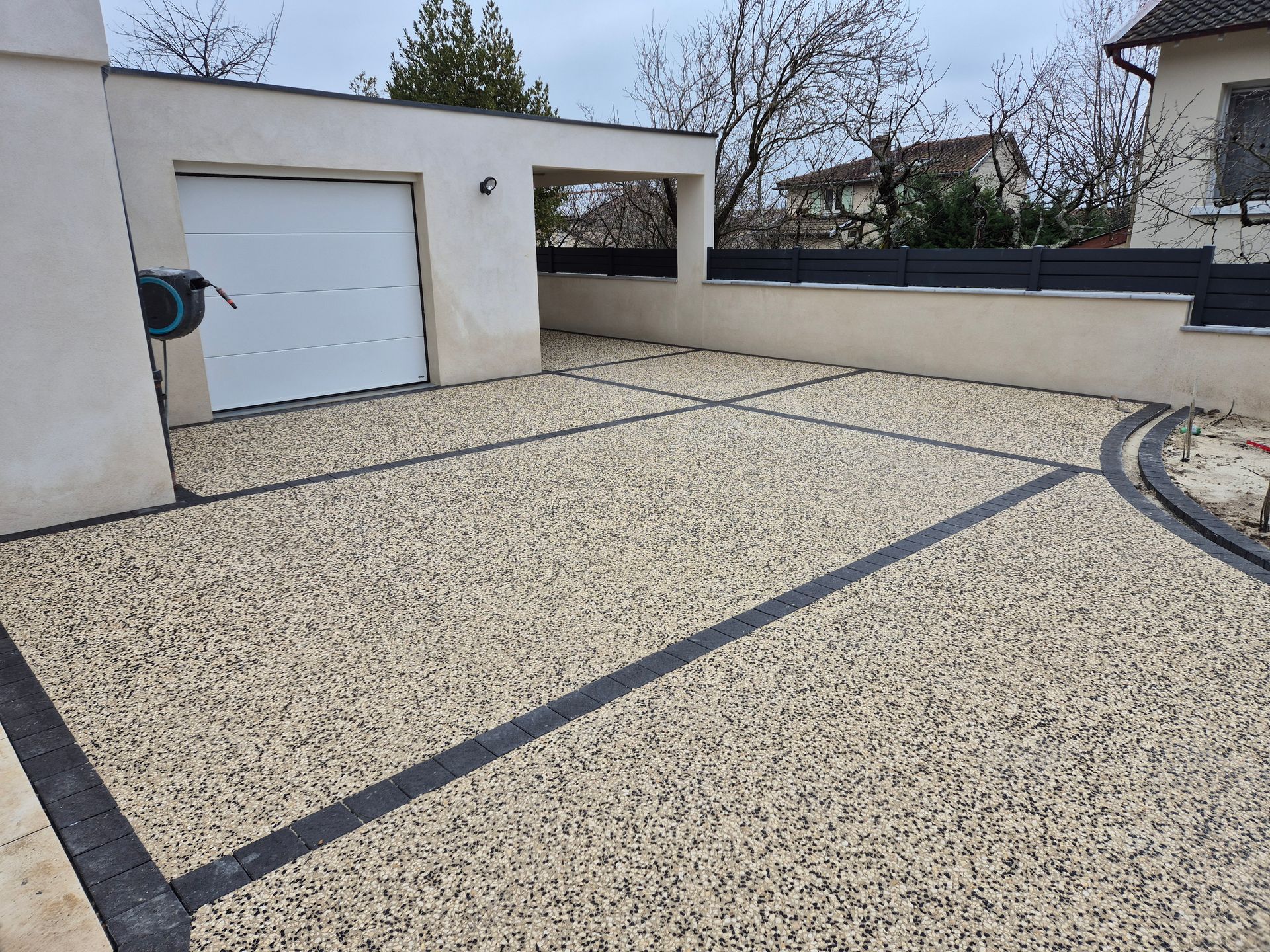 Allée de gravier bordée de lignes sombres, jouxtant un garage et un mur en stuc sous un ciel couvert.