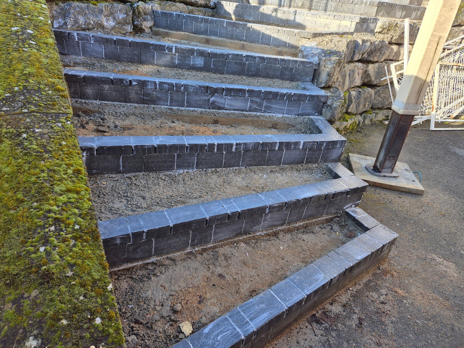 Escaliers extérieurs en pierre avec une rampe en métal, le long d'un chemin de terre et d'un mur recouvert de mousse