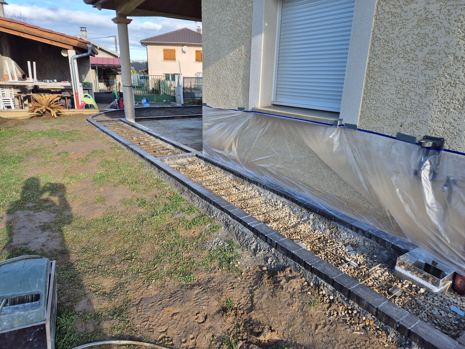 Extérieur d'une maison avec tranchée de gravier, fondations apparentes et fenêtre recouverte d'une bâche en plastique.